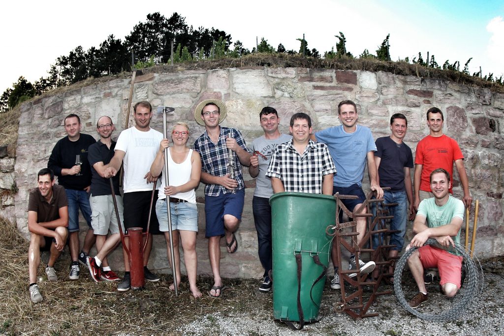Zu sehen sind 12 Personen, die gemeinsam vor einer Mauer stehen. Sie bilden ein Team und halten jeweils Utensilien für den Wein-Anbau in der Hand. Im Hintergrund ist ein Weingut zu sehen.