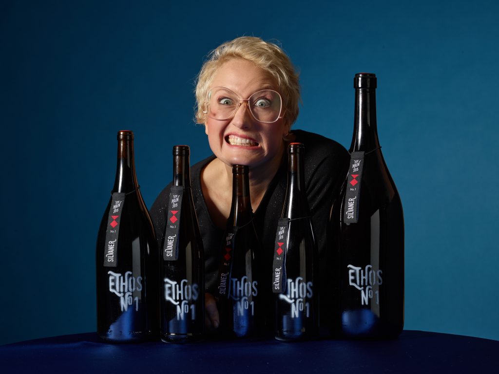 Die blonde Winzerin Ilonka steht hinter 5 Ethos-Weinschlafen und legt Ihren Kopf auf einer Weinflasche ab. Die Flaschen stehen auf einem bauen Tisch, auch der Hintergrund ist blau.