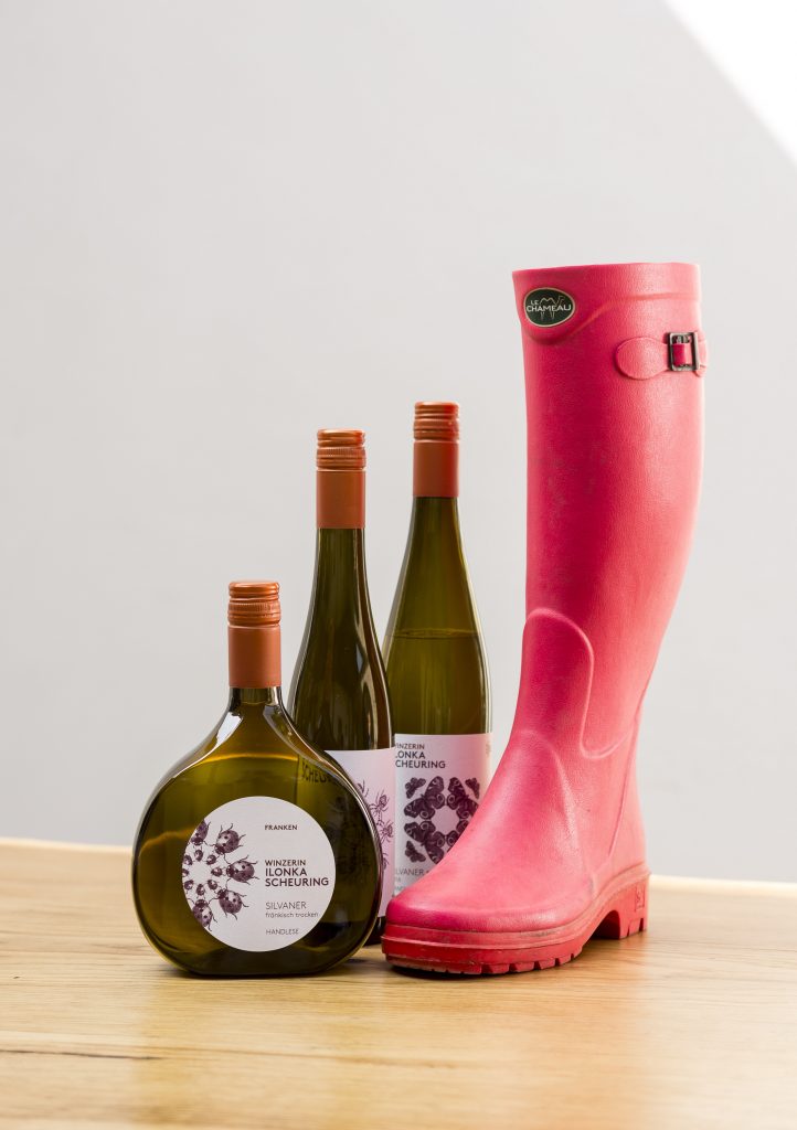 Auf dem Bild sind 3 verschiedene Weinflaschen neben einem pinken Gummistiefel zu sehen. Weinflaschen und Stiefel stehen auf einem hellen Tisch, im Hintergrund befindet sich eine helle Wand.